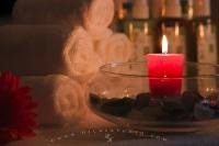 A red candle spreads its relaxing glow and light as it sits in a glass bowl partially filled with sea glass. In the background can be seen a pyramid of rolled towels and some beauty products ready to be used at this spa.
