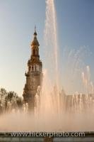 In the Spanish city of Sevilla in Andalusia, a water fountain glistens in the lighting of the sunset in the Plaza de Espana in Parque Maria Luisa with a historic tower decorating the backdrop.
