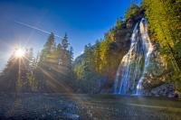 Virgin Falls, a beautiful waterfall set deep in the rainforest on the West Coast of Vancouver Island, glistens in the afternoon sun that beams from a clear blue sky through the trees.