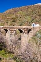 An arched bridge extends across a river next to the town of Valor, Las Alpujarras, which is on the fringes of the Parque Natural de Sierra Nevada, located in the Province of Granada in gorgeous Andalusia, Spain.