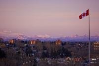 A view of the Rocky Mountains of Alberta tinged a shade of pink by the sunset as seen from the city of Calgary with a Canadian Flag flying high in the cool breeze.