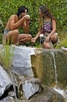 Maori people, a maiden and a warrior, crouch beside a waterfall while drinking from a conch shell at the Wairakei Terraces in New Zealand.
