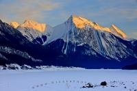 Medicine Lake is covered in snow during winter, while the sunset glows on the snow capped mountain peaks which surround it and the beautiful scenery of Jasper National Park in Alberta, Canada.