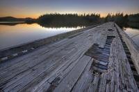 An old and worn out wooden bridge spans the entrance to Clayoquot Arm of Kennedy Lake and leads into a silhouetted scenic background as sunset lights the horizon at the close of another day.