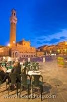 As people sit outside a cafe enjoying the ambiance and architecture of Piazza Del Campo, the Torre del Mangia and Palazzo Pubblico are lit up as dusk falls on the historic old town district in Siena; a UNESCO World Heritage Site.