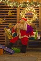 A mannequin dressed up like santa - placed on a park bench and surrounded by gifts and sparkling lights forms part of a christmas display outside a house.