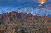 The moon shines brightly behind a series of cloud formations above the Sierra Nevada Mountain range and above the surreal scenery and rock formations of the Alabama Hills at twilight.