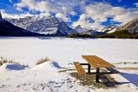 A picnic table which sits along the shore of the snow covered Upper Kananaskis Lake is surrounded by beautiful winter scenery. The Kananaskis mountain range is a beautiful backdrop to the lake.