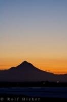 The volcano peak of Mt. Taranaki on the North Island of New Zealand is silhouetted by the yellow stretch of sky at sunset.
